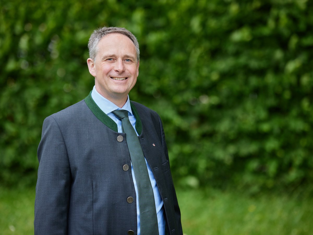 Lächelnder Mann im Trachtenjanker mit grüner Krawatte vor unscharfem Grün.