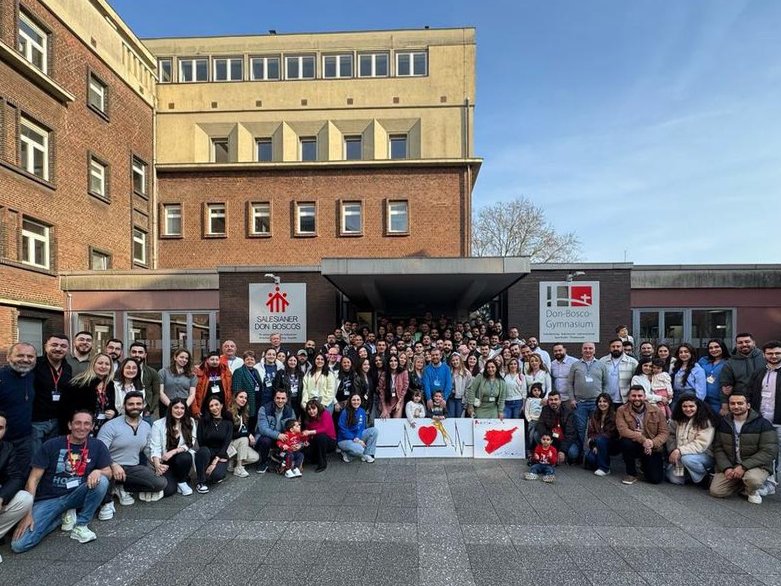 Eine große Gruppe von Menschen posiert vor dem Don-Bosco-Gymnasium mit Plakaten, die ein Herz und Landkarten zeigen.