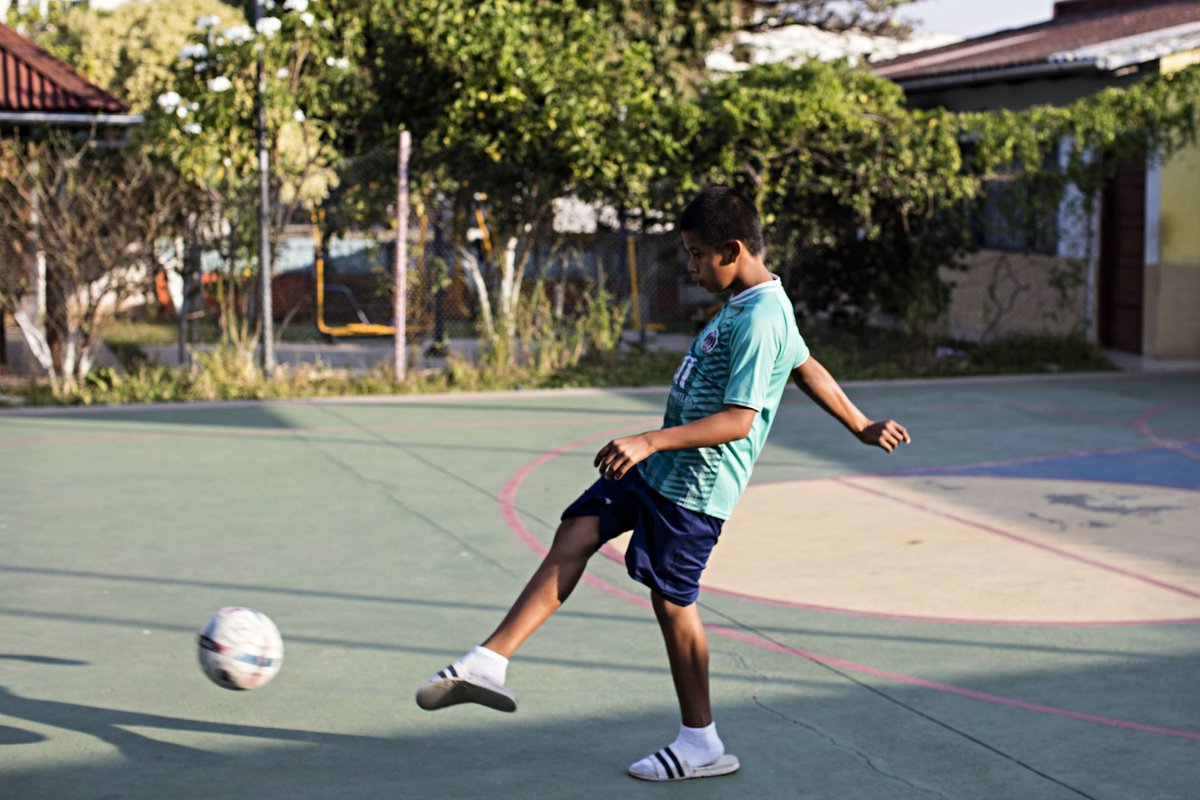 Ein junger Junge in einem türkisen Trikot kickt einen Fußball auf einem grünen Outdoor-Platz, Bäume im Hintergrund.
