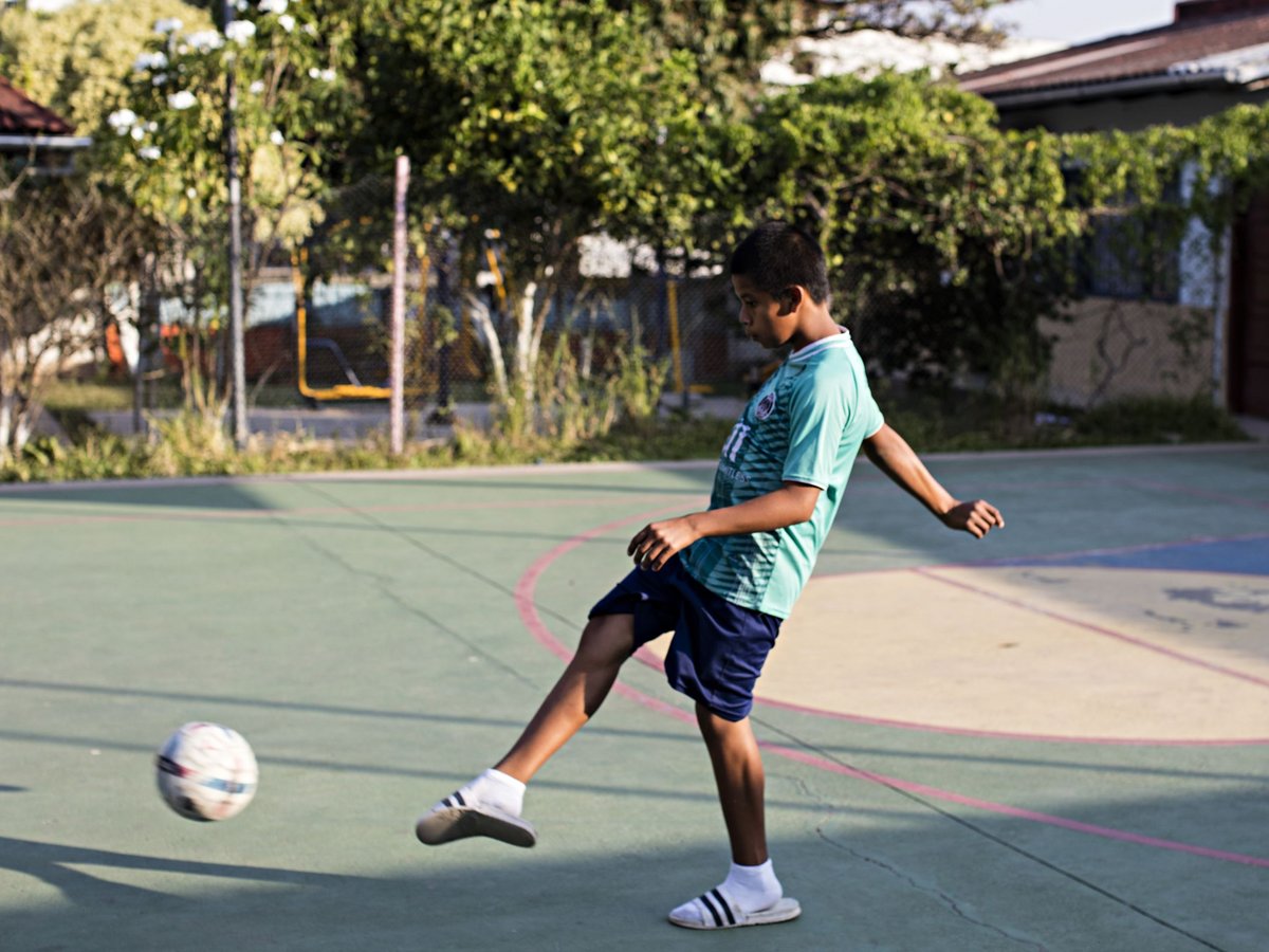 Ein junger Junge in einem türkisen Trikot kickt einen Fußball auf einem grünen Outdoor-Platz, Bäume im Hintergrund.