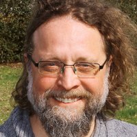 Porträt eines lächelnden Mannes mit Brille, langen braunen Haaren und grauem Bart, der einen grauen Strickpullover trägt.