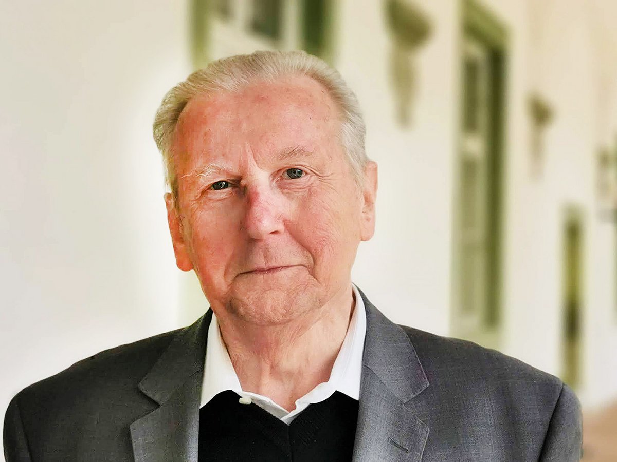 Porträt eines älteren Mannes mit grauem Haar, Anzugjacke und weißem Hemd, der direkt in die Kamera blickt.