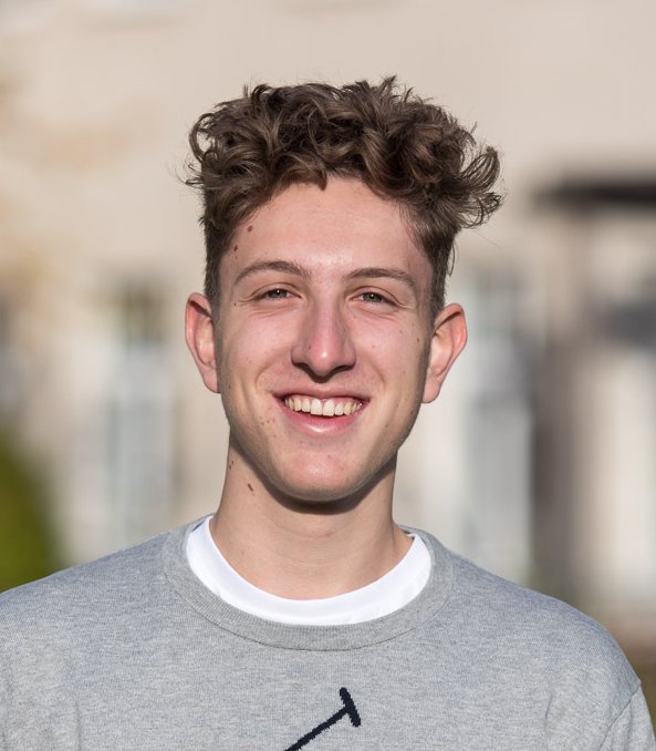 Ein lächelnder junger Mann mit lockigem Haar, der ein graues Sweatshirt mit großem Ralph Lauren Polo-Logo trägt. Im Freien.