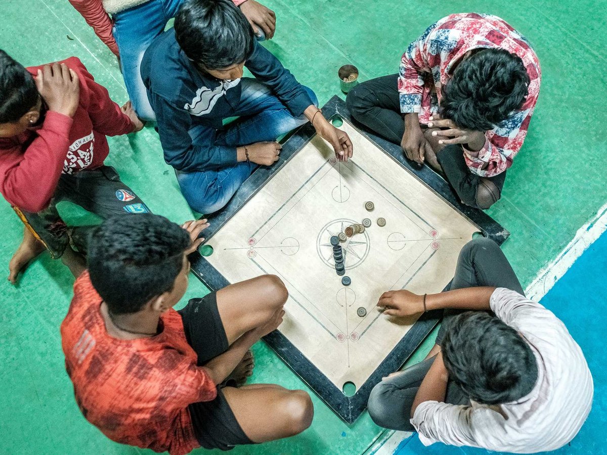 Vogelperspektive: Mehrere junge Männer spielen Carrom auf einem Brett auf grünem Boden; einer macht einen Zug.