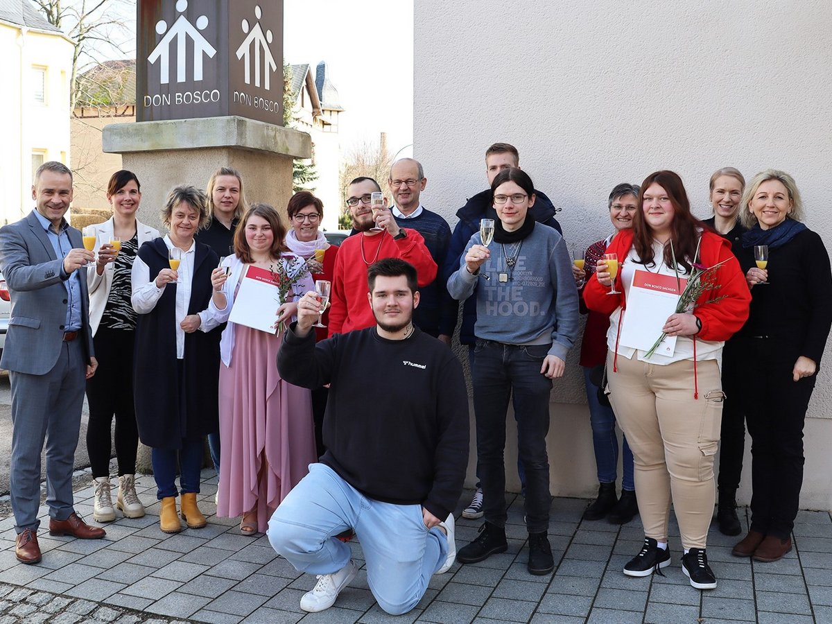 Eine Gruppe von etwa 15 Personen feiert vor einem Don Bosco Schild, viele halten Sektgläser, einige Dokumente und Blumen.