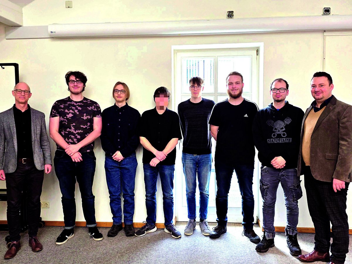 Acht Männer, einer mit verpixeltem Gesicht, stehen in einer Reihe in einem hell beleuchteten Raum.