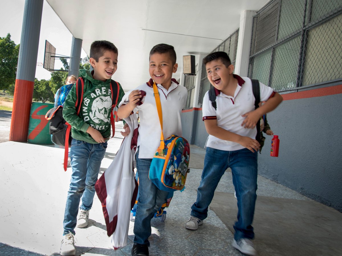 Drei Schulkinder laufen lachend mit Rucksäcken durch einen Gang auf einem Schulhof. Sie wirken ausgelassen und glücklich.