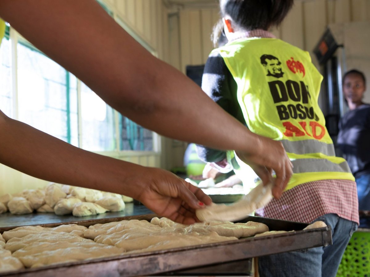 Personen bereiten Teig in einer Küche zu, eine trägt eine gelbe Weste mit „DON BOSCO AID“, legen Teiglinge auf Bleche.