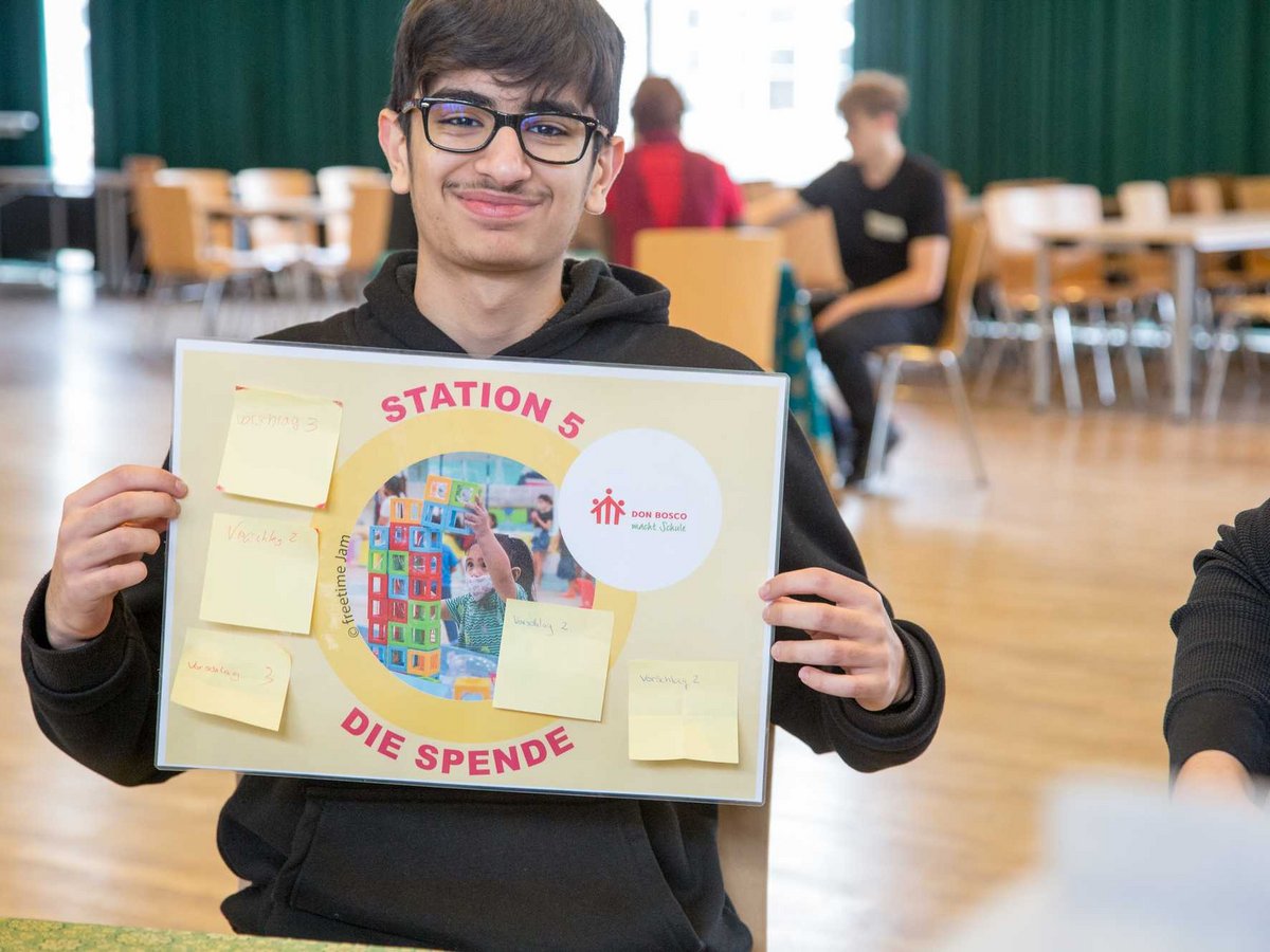 Lächelnder junger Mann mit Brille hält ein Schild „STATION 5 DIE SPENDE“ mit Don Bosco Logo und Bild eines spielenden Kindes.