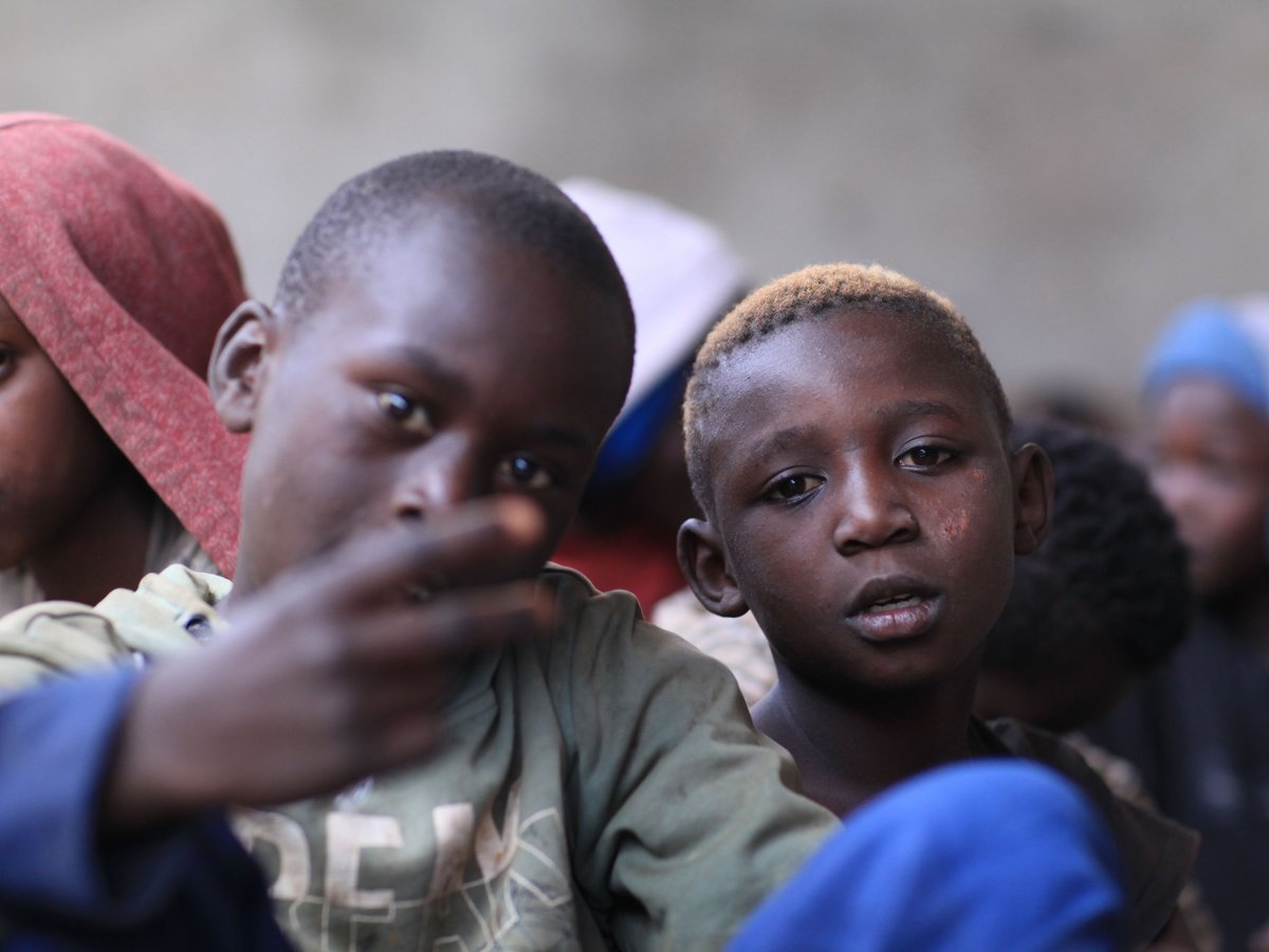 Nahaufnahme von zwei dunkelhäutigen Kindern; eines gestikuliert mit Fingern, das andere mit kurzen hellen Haaren blickt direkt an.