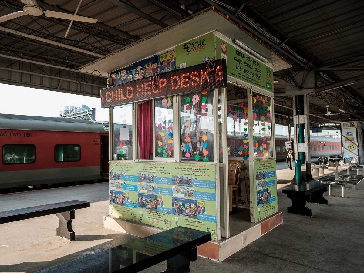 Ein farbenfroher 'Child Help Desk' Stand an einem indischen Bahnhof mit einem roten Zug und Bänken.