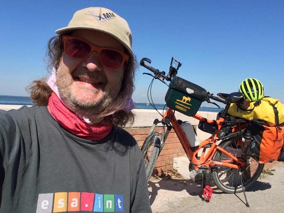 Ein lächelnder Mann mit Sonnenbrille und Kappe macht ein Selfie mit seinem beladenen orangefarbenen Touren-E-Bike am Meer.