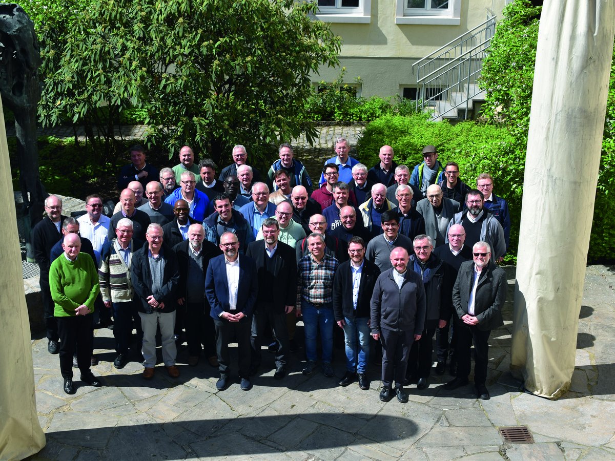 Große Gruppe Männer, möglicherweise Geistliche, lächeln für ein formelles Gruppenfoto im Freien an einem sonnigen Tag.