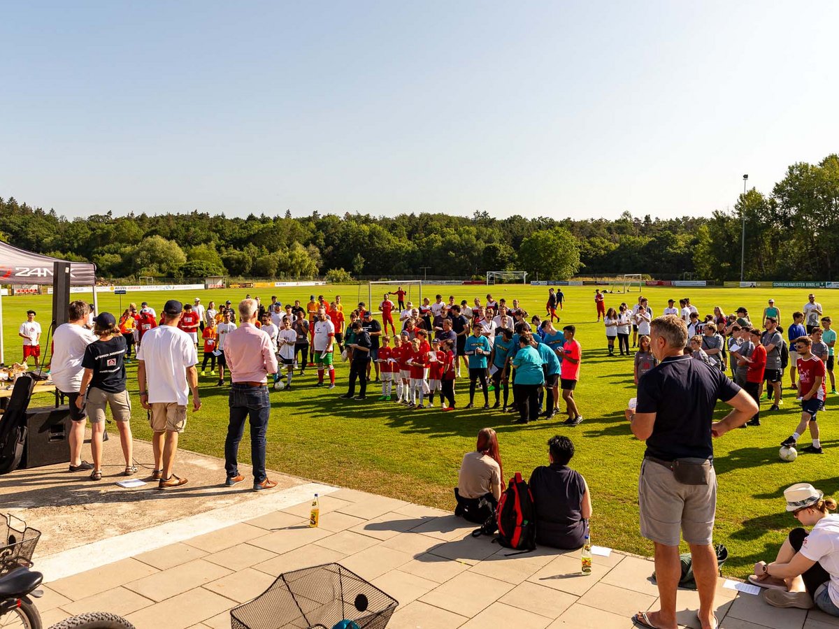 Viele Kinder und Erwachsene versammeln sich auf einem sonnigen grünen Fußballfeld für ein Sportevent oder Camp, mit einem Zelt daneben.