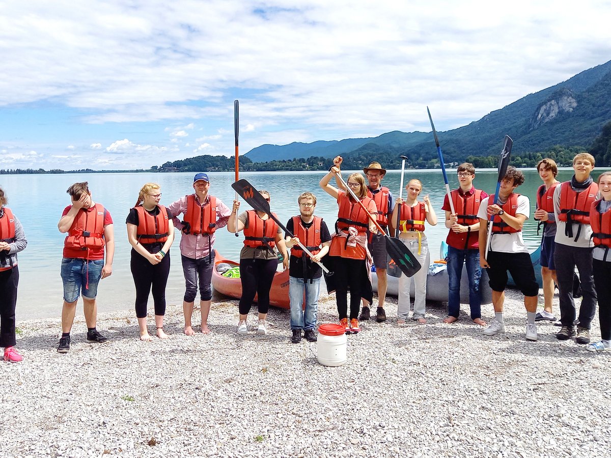 Eine Gruppe Jugendlicher und Erwachsener mit roten Schwimmwesten und Paddeln steht an einem Kiesstrand vor einem See und Bergen.