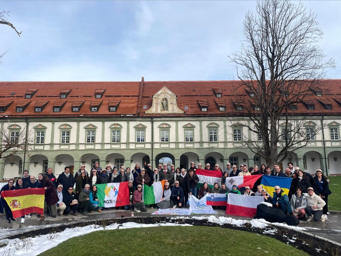 Gruppe von Menschen mit Nationalflaggen posiert vor einem historischen Gebäude mit rotem Dach und Schnee auf dem Boden.