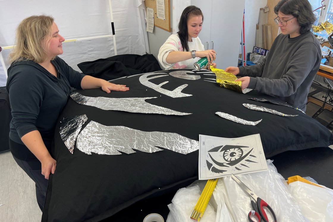 Drei Personen basteln ein Augendesign auf schwarzem Stoff mit Silber- und Goldfolie.