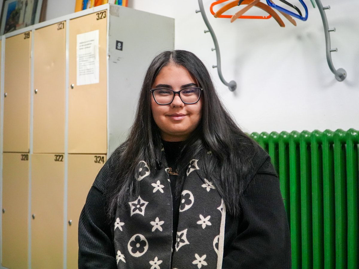 Junge Frau mit langen dunklen Haaren und Brille lächelt in einem Raum mit Spinden, grünem Heizkörper und Kleiderbügeln.