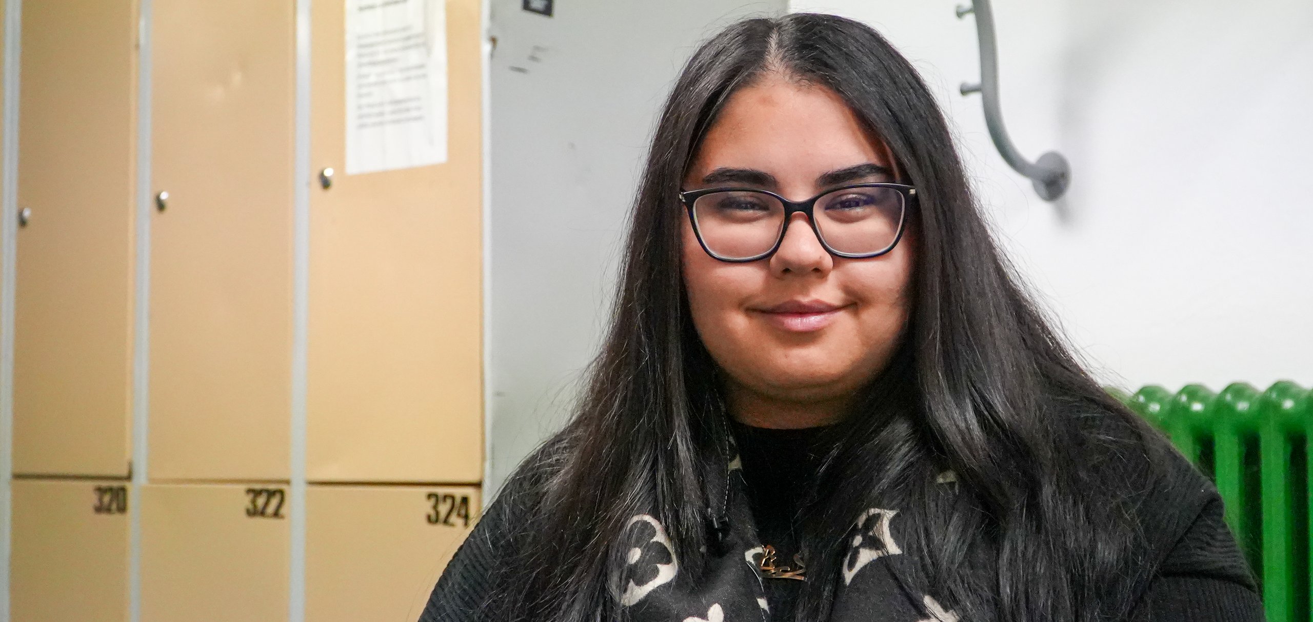 Junge Frau mit langen dunklen Haaren und Brille lächelt in einem Raum mit Spinden, grünem Heizkörper und Kleiderbügeln.