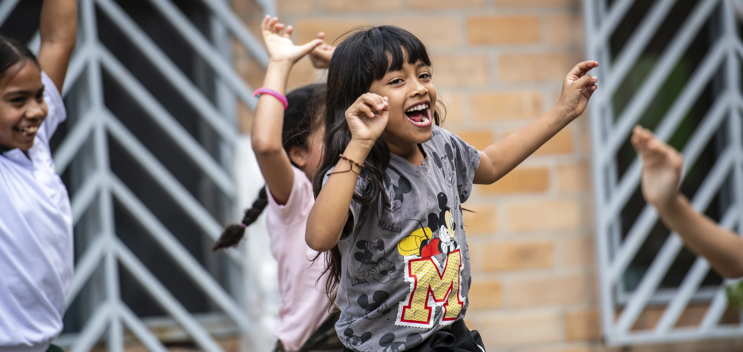 Lächelndes Mädchen im Mickey-Maus-Shirt tanzt enthusiastisch mit anderen Kindern im Freien vor einer Ziegelwand.