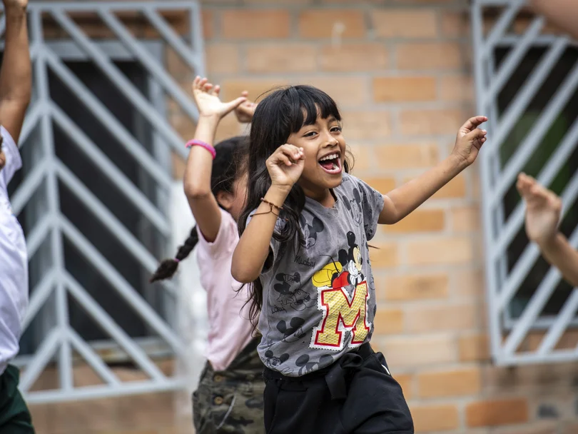 Lächelndes Mädchen im Mickey-Maus-Shirt tanzt enthusiastisch mit anderen Kindern im Freien vor einer Ziegelwand.