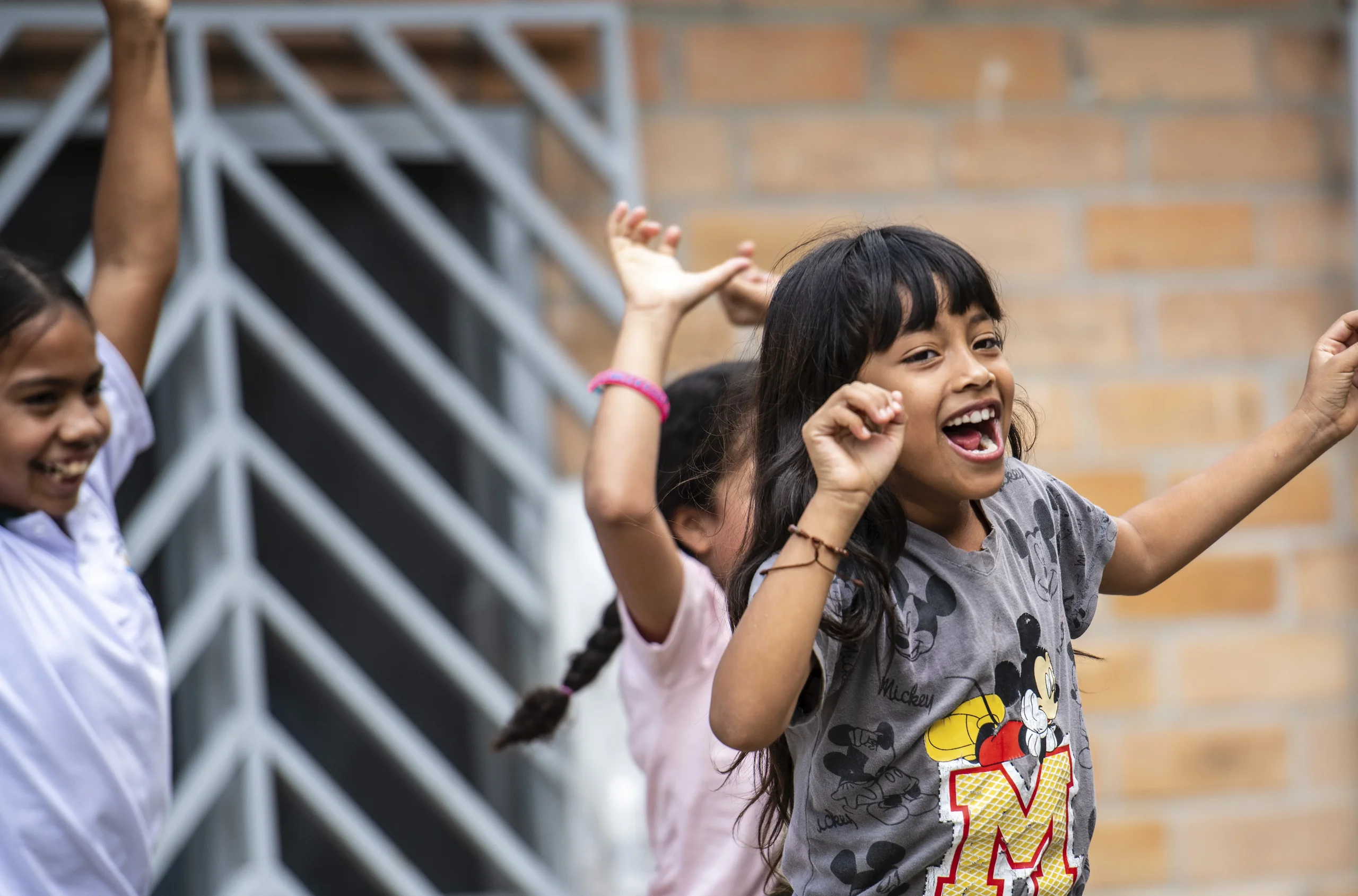 Lächelndes Mädchen im Mickey-Maus-Shirt tanzt enthusiastisch mit anderen Kindern im Freien vor einer Ziegelwand.