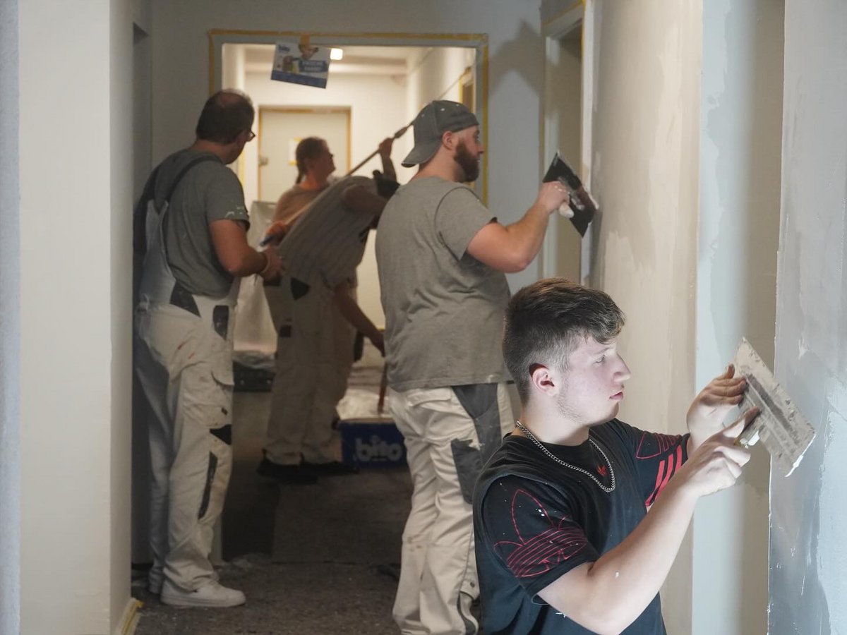Ein junger Mann verputzt eine Wand mit einer Kelle, während im Hintergrund weitere Handwerker arbeiten.