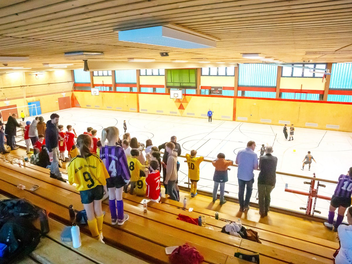 Hallensoccer-Spiel in einer Sporthalle, Kinder spielen, Zuschauer beobachten von den Holztribünen.