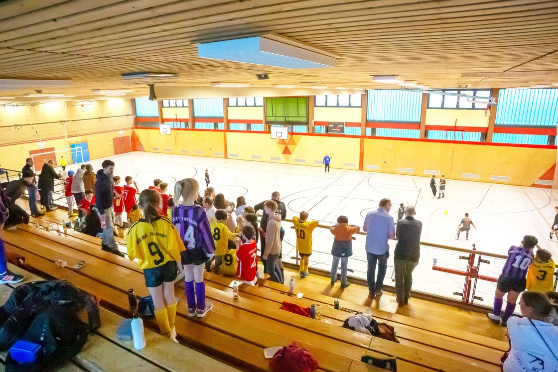 Hallensoccer-Spiel in einer Sporthalle, Kinder spielen, Zuschauer beobachten von den Holztribünen.