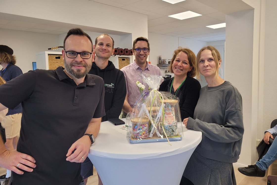 Eine Gruppe von fünf lächelnden Personen, drei Männer und zwei Frauen, steht um einen Stehtisch mit einem Geschenkkorb.