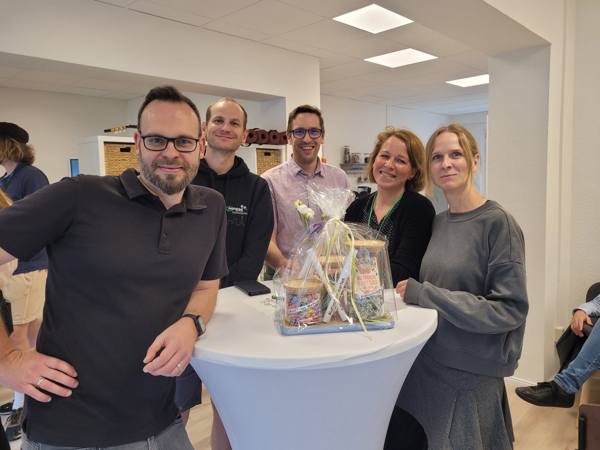 Eine Gruppe von fünf lächelnden Personen, drei Männer und zwei Frauen, steht um einen Stehtisch mit einem Geschenkkorb.