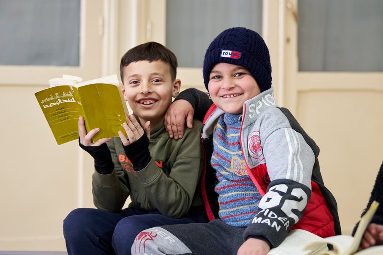 Zwei Jungen lächeln glücklich in die Kamera, einer hält ein Buch mit arabischer Schrift und der andere trägt eine blaue Mütze.