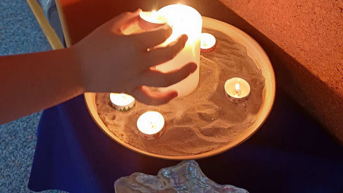 Kinderhand zündet Teelicht an brennender Kerze an, daneben brennende Teelichte in Schale mit Sand