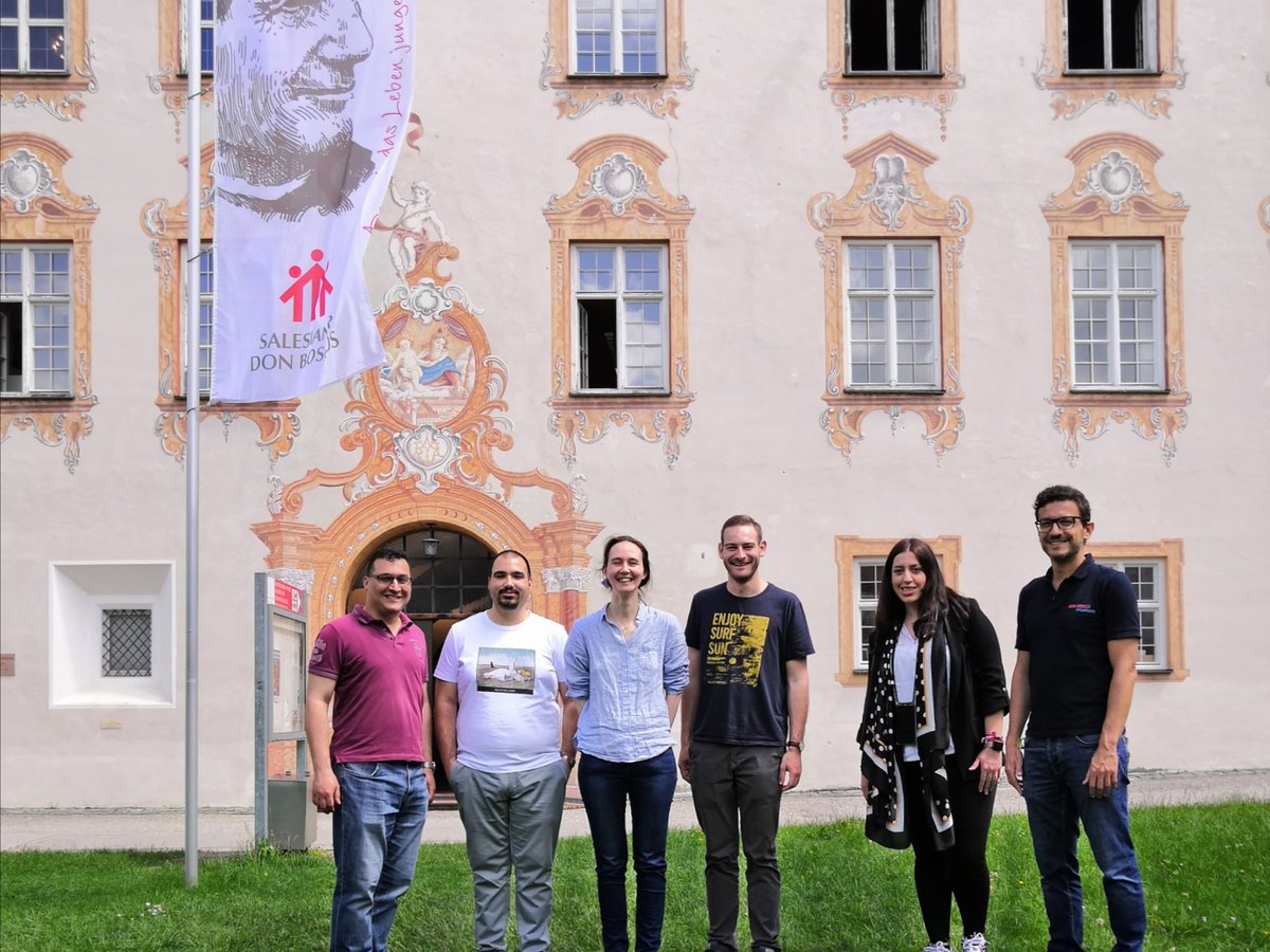Sechs Personen stehen auf einer Wiese vor einem historischen Gebäude mit einem Don Bosco Banner.