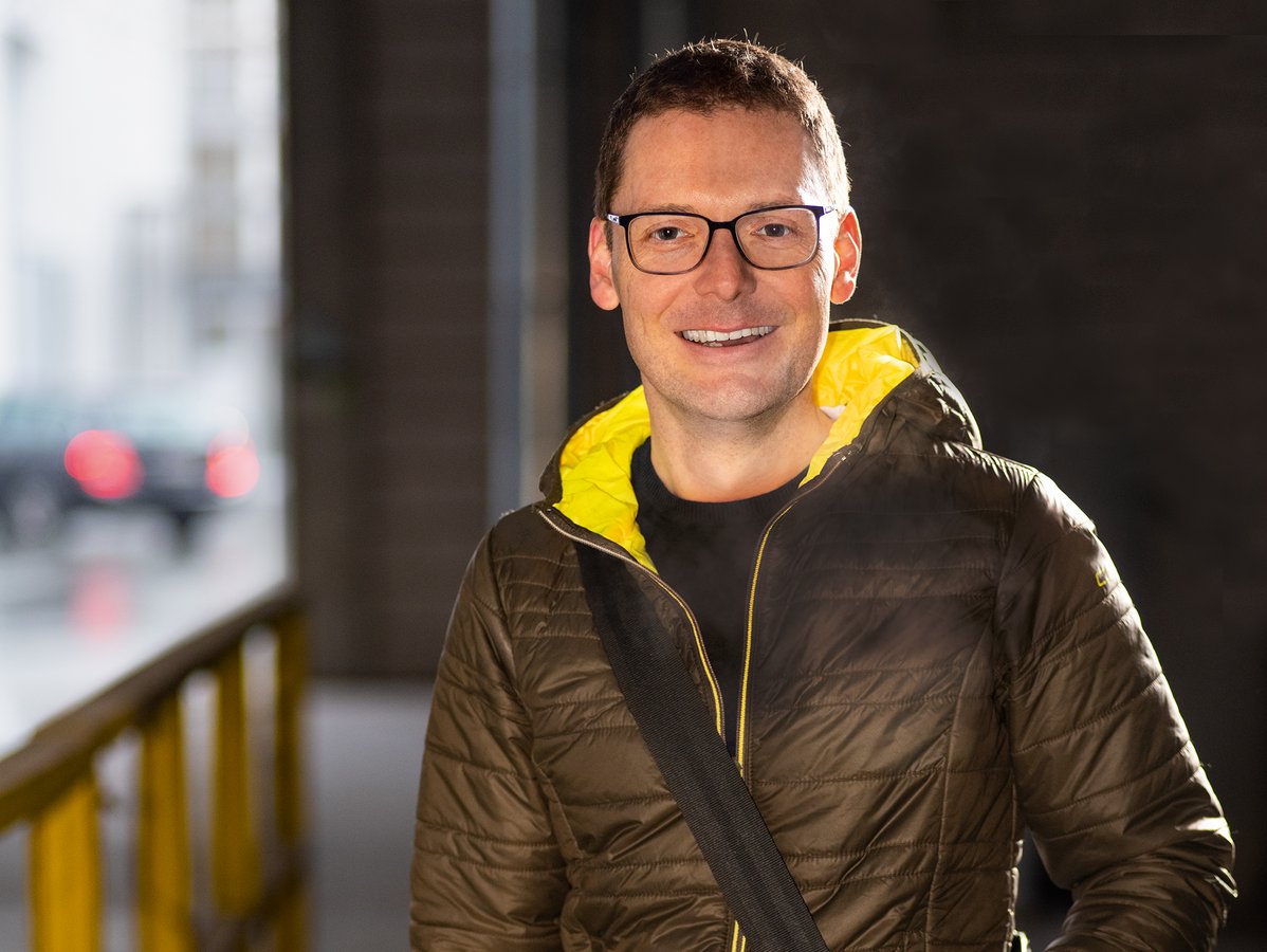 Ein lächelnder Mann in grüner Jacke und Brille, eine Umhängetasche tragend, steht in einer Unterführung.