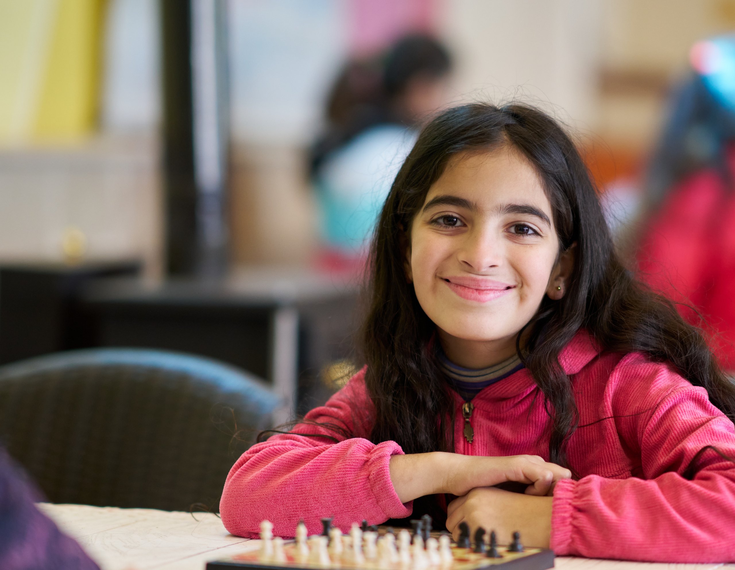 Lächelndes junges Mädchen mit langen dunklen Haaren in einem pinken Hoodie sitzt an einem Tisch mit einem Schachbrett.