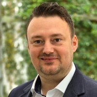 Ein freundlich lächelnder Mann in dunklem Anzug und weißem Hemd vor einem unscharfen grünen Hintergrund.