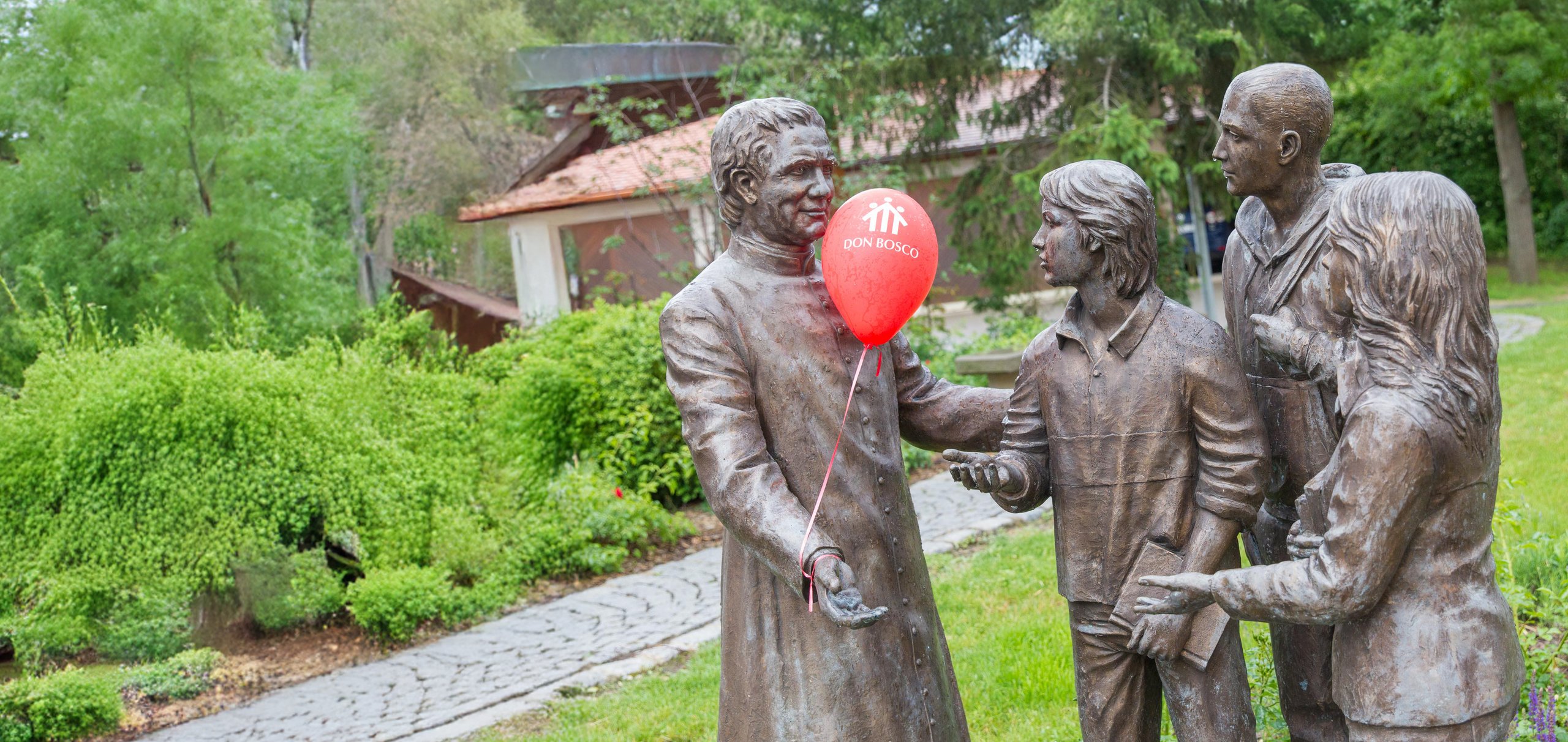 Bronzestatue von Don Bosco mit rotem "Don Bosco"-Ballon, im Gespräch mit drei Jugendstatuen in einem grünen Park.