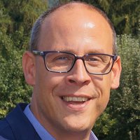 Lächelnder Mann mit Brille, blauem Sakko und hellblauem Tupfenhemd, draußen vor verschwommenem grünem Laub.