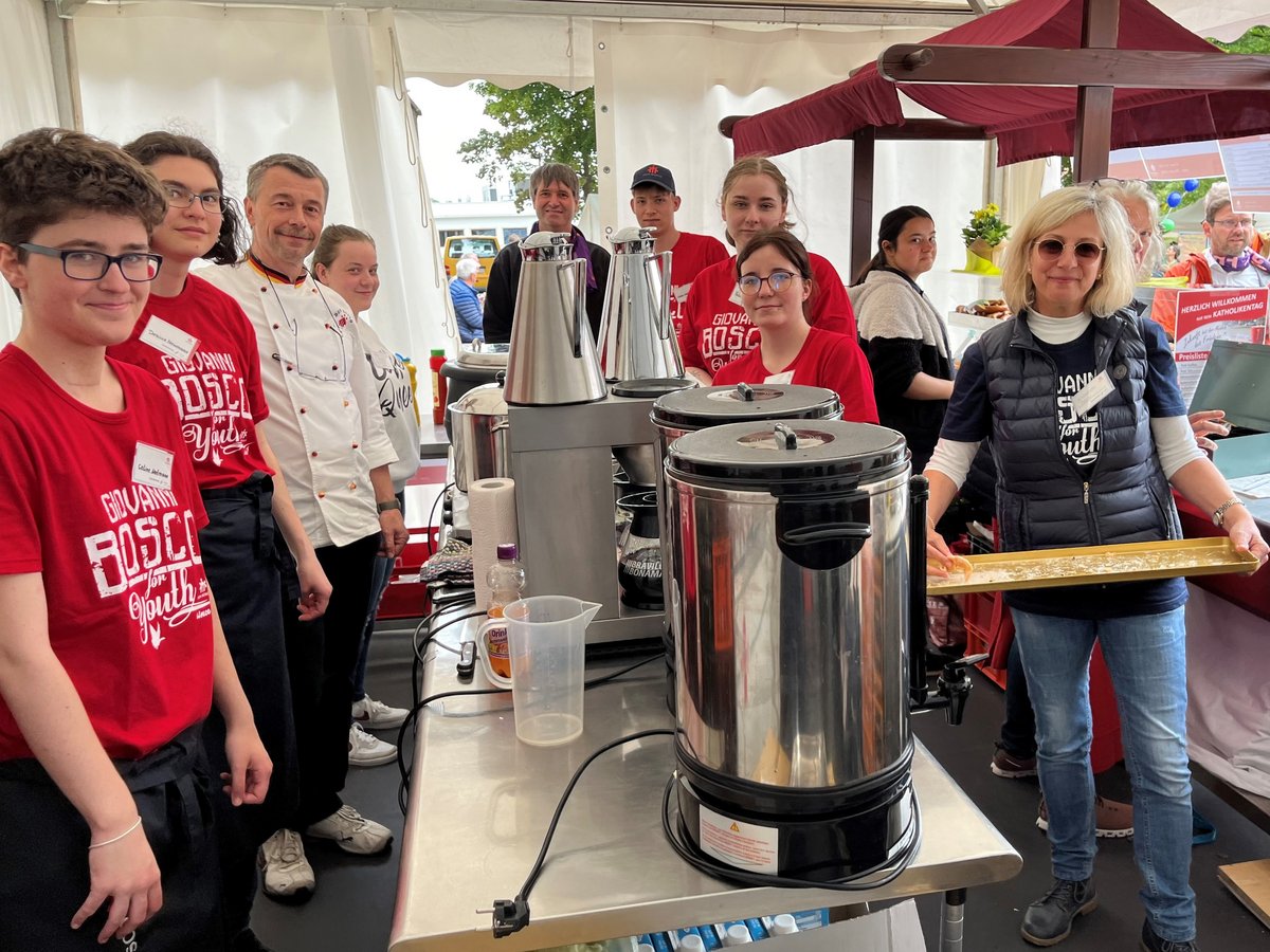 Freiwillige in roten 'Giovanni Bosco for Youth'-Shirts und andere lächeln hinter einem Verkaufsstand mit Getränkeautomaten bei einem Event.