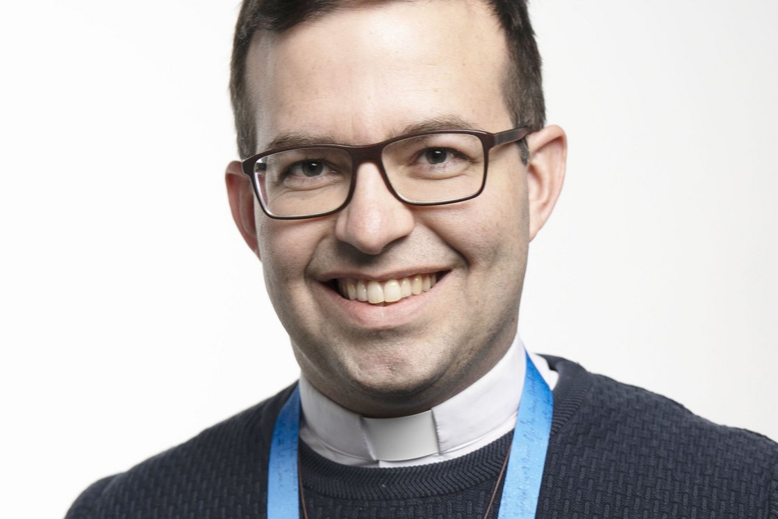 Lächelnder Mann mit dunklem Haar, Brille, Klerikerkragen, dunklem Pullover und blauem Schlüsselband blickt in die Kamera.