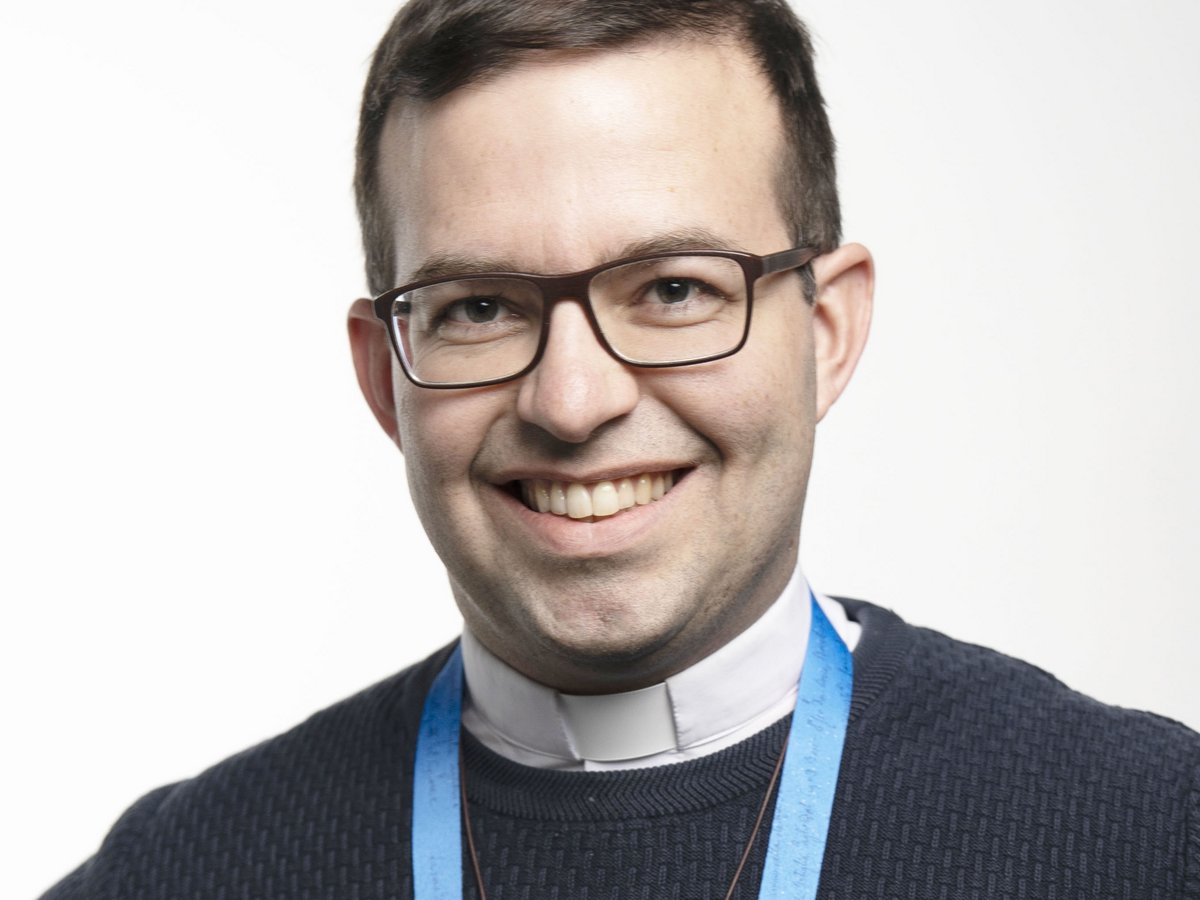 Lächelnder Mann mit dunklem Haar, Brille, Klerikerkragen, dunklem Pullover und blauem Schlüsselband blickt in die Kamera.