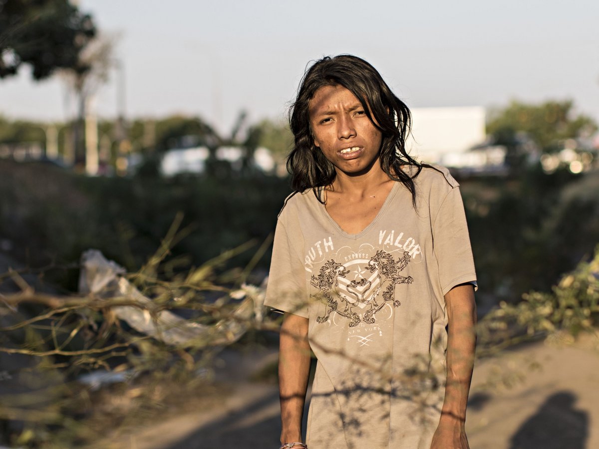 Eine junge Person mit langen dunklen Haaren und dunkler Haut blickt ernst in die Kamera, mit gesprenkeltem Sonnenlicht und Schatten im Gesicht.