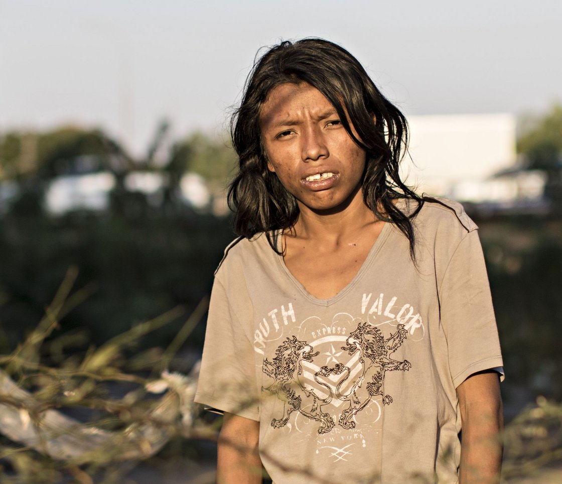 Eine junge Person mit langen dunklen Haaren und dunkler Haut blickt ernst in die Kamera, mit gesprenkeltem Sonnenlicht und Schatten im Gesicht.
