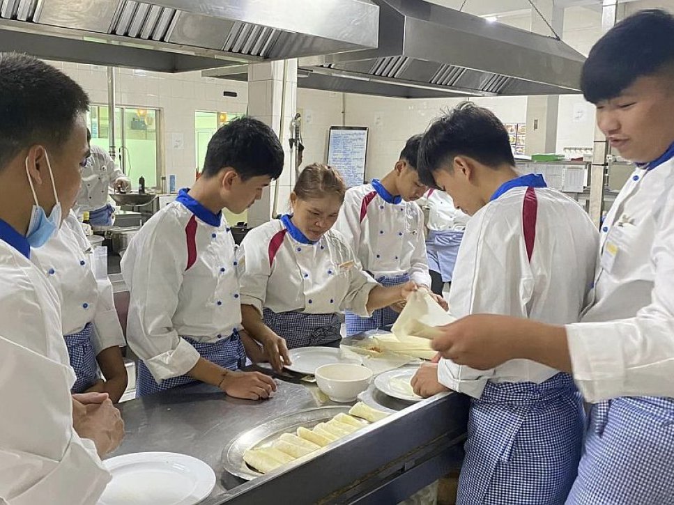 Kochschüler in Uniform bereiten unter Anleitung Speisen, wahrscheinlich Frühlingsrollen, in einer Küche zu.