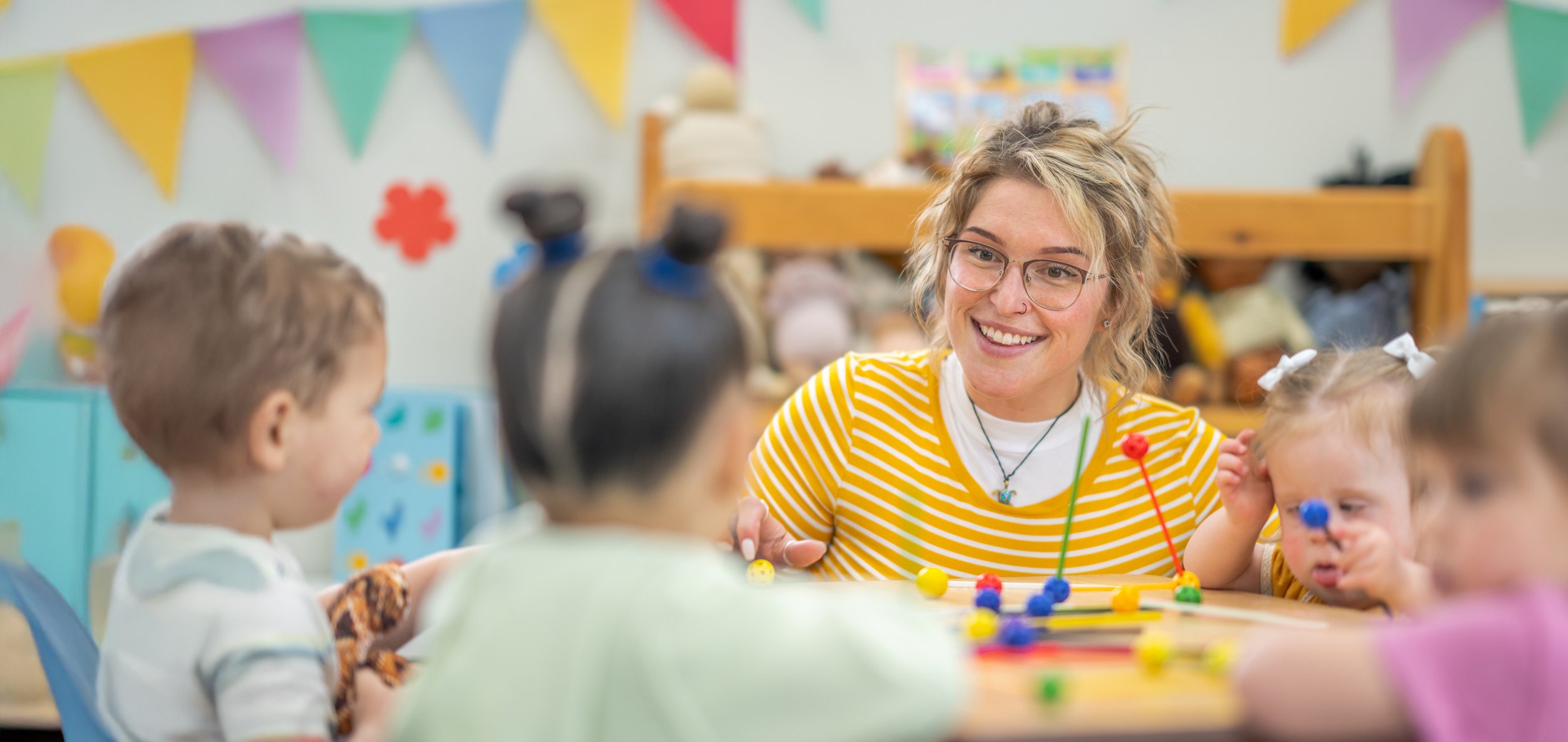 Eine lächelnde Erzieherin mit Brille und blonden Haaren sitzt mit Kleinkindern an einem Tisch, die mit bunten Spielsachen spielen.