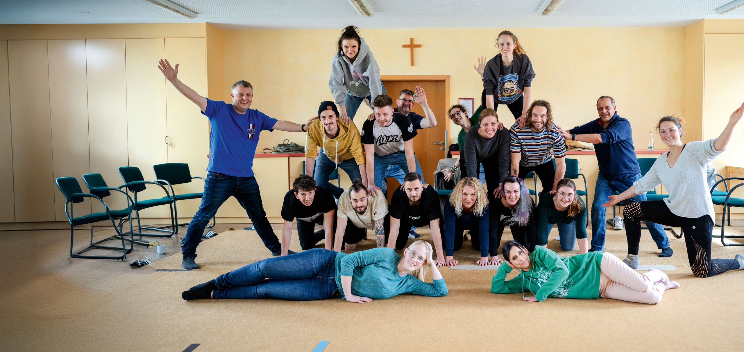 Eine große Gruppe lächelnder Menschen bildet eine Menschenpyramide in einem hellen Raum.