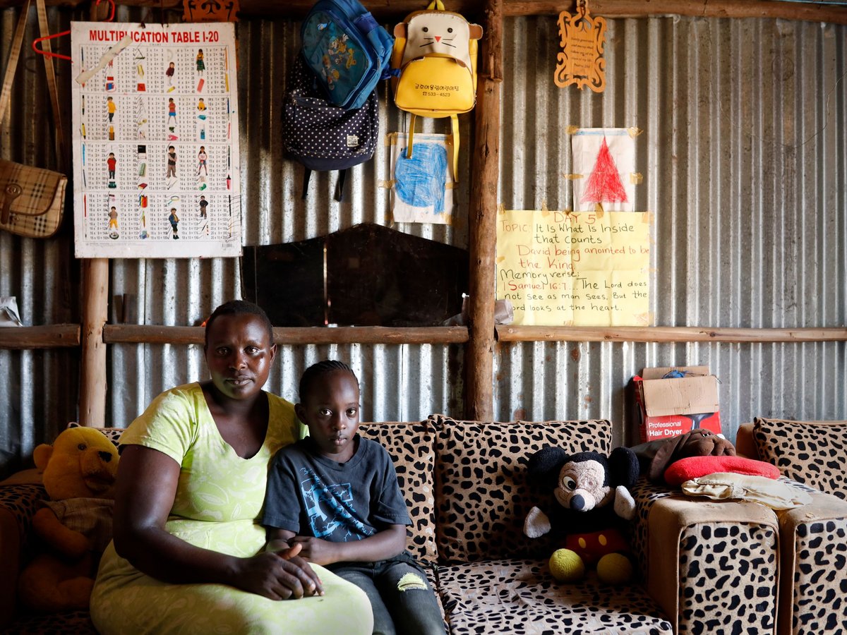 Mutter und Kind sitzen auf einem Leopardenmuster-Sofa in einem Raum mit Wellblechwänden und Schultafeln.