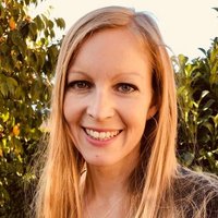 Lächelnde Frau mit langen blonden Haaren, hellen Augen und einer Halskette, vor einem verschwommenen grünen Hintergrund im Freien.