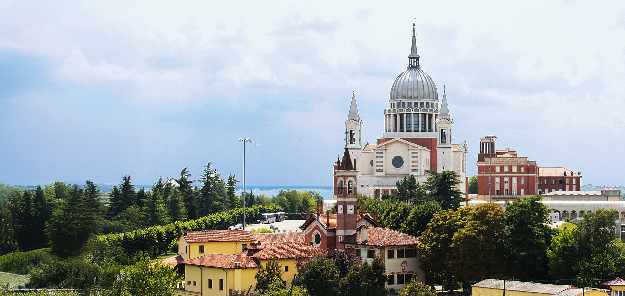 Santuario Don Bosco, eine große Kuppelkirche auf einem Hügel, umgeben von Bäumen und Gebäuden unter bewölktem Himmel.